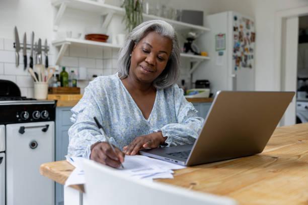 Mature woman at home taking classes online using her laptop Mature black woman at home taking classes online using her laptop and writing some notes virtual office stock pictures, royalty-free photos & images